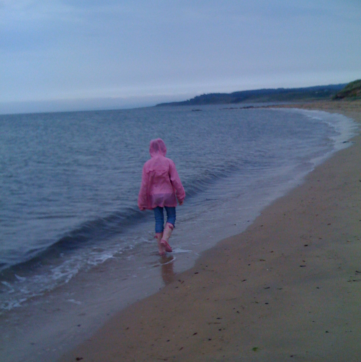 A young person dressed in a pink waterproof and wellies walking along the beach in the wash of the waves.