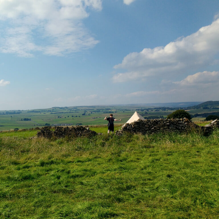 A grassy hill with views over English Countryside. There is a figure jumping over a fallen down stone wall facing away from the camera. A old fashioned tent is visible close by. It's very sunny and there are clouds in the sky.