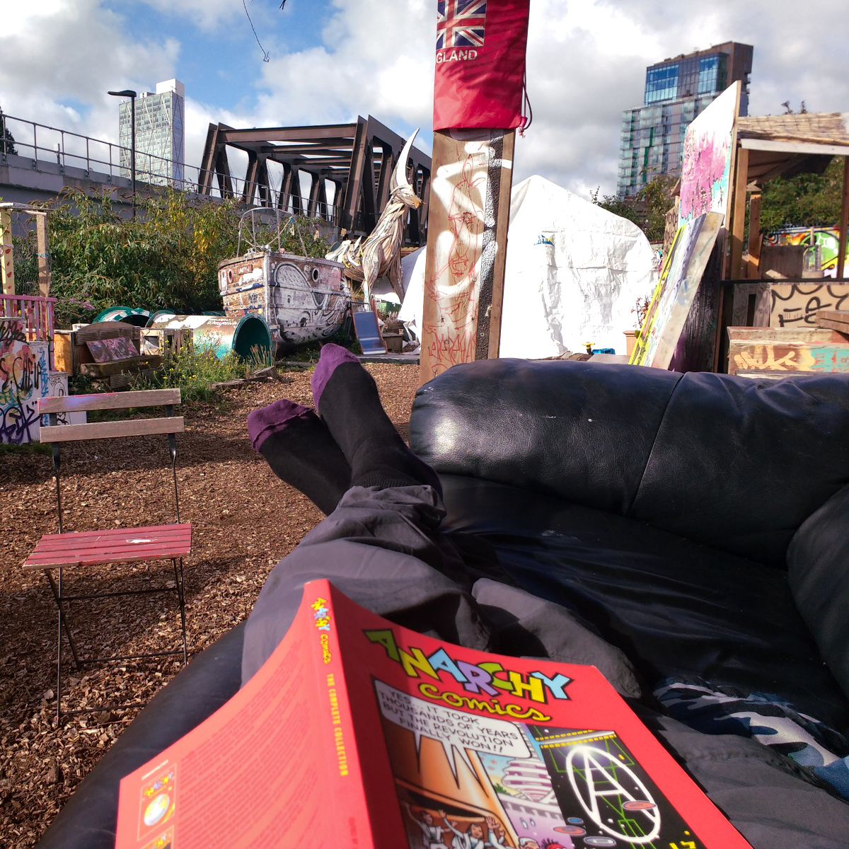 POV photo of a persons legs lying on a sofa outside in some sort of cool punk hacker garden with loads of structures made of wood and graffiti. Tower blocks are in the background. The person is reading Anarchy Comics.