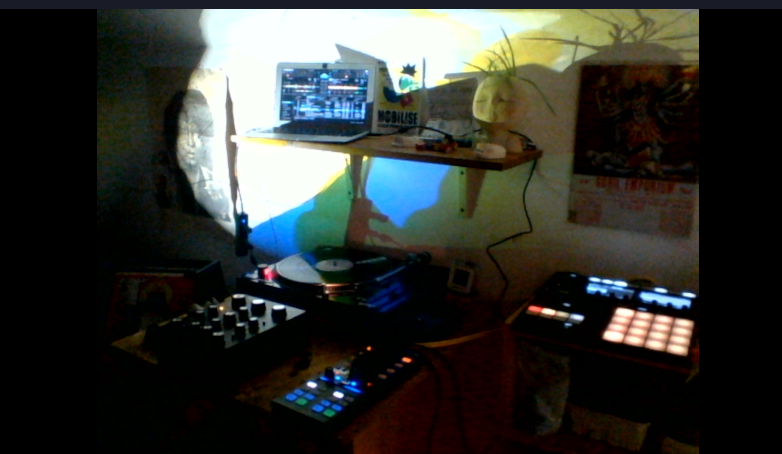 Photo of my DJ area. It's dark with 60s psychedelic lighting. Visible is a Maschine+, a mixer and turntable and a macbook running Traktor. There's loads of weird and curious objects knocking around.